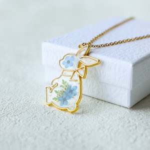 May include: Gold necklace featuring a resin pendant shaped like a rabbit. The pendant is filled with dried blue flowers.