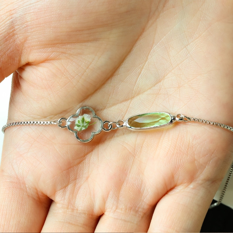 May include: A delicate silver chain bracelet with a clover-shaped charm containing a pressed flower and a light green oval-shaped charm.