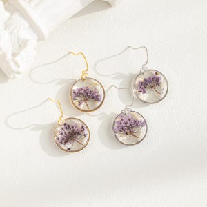 May include: A pair of gold and silver earrings with delicate purple flower accents encased in clear resin. The earrings feature a simple, elegant design.
