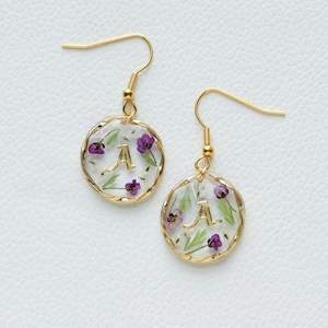 May include: Gold earrings with a clear resin circle containing pressed purple and white flowers and a gold letter 'A'.