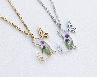 Puede incluir: Dos delicados collares con un colgante de gato con una mariposa y flores prensadas. El gato está contorneado en metal dorado y plateado.