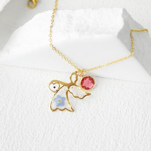 May include: Gold necklace with a delicate angel charm featuring a blue flower and a red gemstone. The angel charm is suspended from a gold chain.