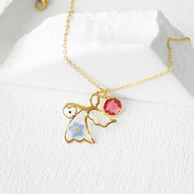 May include: Gold necklace with a delicate angel charm featuring a blue flower and a red gemstone. The angel charm is suspended from a gold chain.