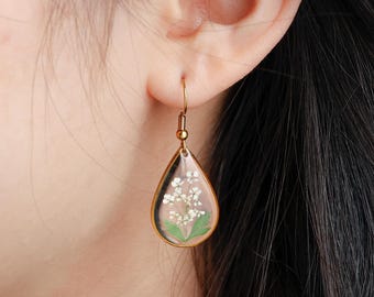 May include: Gold teardrop earrings with a clear resin design featuring a small white flower and green leaves.