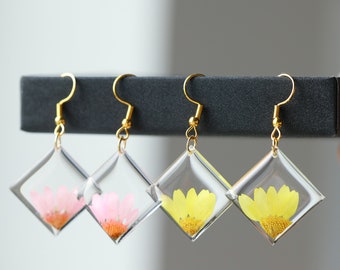Könnte beinhalten: Drei Paar Ohrringe mit goldenen Haken und klaren quadratischen Harzfassungen. Jedes Paar zeigt eine andere gepresste Blume: rosa, gelb und gelb.