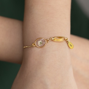Könnte beinhalten: Ein goldenes Armband mit einer zarten Kette, das einen durchsichtigen ovalen Anhänger mit einer gepressten weißen Blume, einen goldenen ovalen Anhänger mit einem durchsichtigen gelben Stein und einen kleinen goldenen Anhänger mit der eingravierten Aufschrift "Life" trägt.