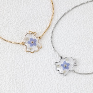 Könnte beinhalten: Zwei zarte Armbänder mit einer goldfarbenen und einer silberfarbenen Kette. Jedes Armband hat einen transparenten, geisterförmigen Anhänger, der eine gepresste blaue Blume und kleine weiße Blütenblätter im Inneren des Materials enthält.