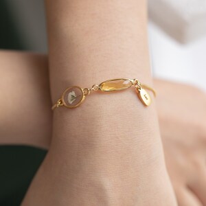Puede incluir: Un brazalete de oro con una cadena delicada, que presenta un peque&ntilde;o dije redondo con una flor prensada, una piedra rectangular de color &aacute;mbar y un peque&ntilde;o dije redondo con la letra "L".