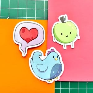 May include: Three colorful stickers on a green and orange surface. The stickers include a red heart in a speech bubble, a green apple with a smiling face, and a blue chicken with a purple comb.