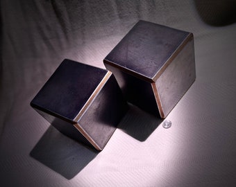May include: Two dark gray metal cubes with a thin gold trim. The cubes are sitting on a white surface.