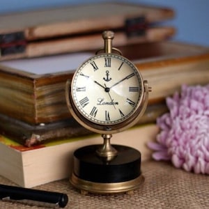 May include: A brass desk clock with a round face and Roman numeral markings. The clock has a black base and an anchor design on the face. The clock face reads "London".