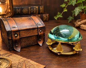 May include: A dome-shaped glass paperweight magnifier with a polished gold-tone metal base featuring decorative fluted columns. The clear, rounded lens sits atop the circular frame, providing a clear view of the surface beneath it.
