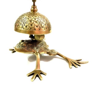 Pode incluir: Uma campainha de porta em forma de sapo de cor dourada com um topo de sino adornado. O sino &eacute; decorado com um padr&atilde;o floral.