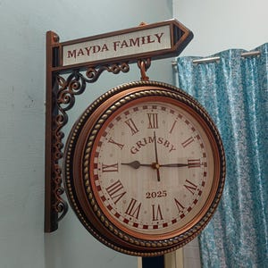 May include: A double-sided wall clock with the text "MAYDA FAMILY" on a sign above. The clock face is cream-coloured with Roman numerals and the word "GRIMSBY". The clock is brown and bronze with decorative scrollwork.