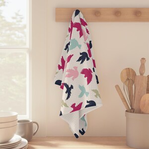May include: A white kitchen towel with a pattern of colorful birds in flight. The birds are in shades of pink, blue, and black. The towel is hanging on a wooden rack.