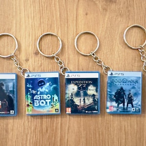 Peut inclure: Cinq porte-clés avec des boîtiers de jeux PS5 miniatures. Les jeux incluent "The Last of Us Part II", "Astro Bot", "Expedition 33", "God of War Ragnarok", et un jeu avec un couple qui s'embrasse. Chaque porte-clés a un anneau argenté.