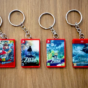 Op de afbeelding: Vier rode sleutelhangers met artwork van videogames. De sleutelhangers hebben zilveren ringen en kettingen. De gamecovers tonen Super Mario Odyssey en The Legend of Zelda: Breath of the Wild.