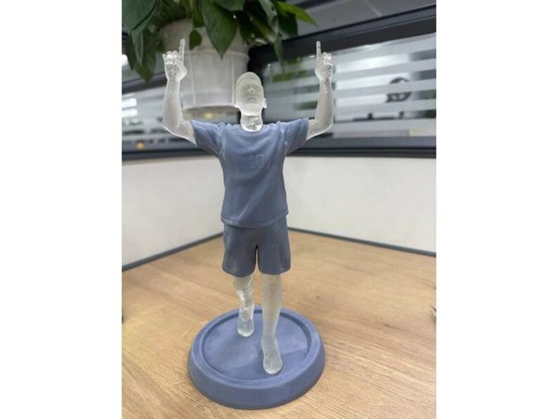 May include: A clear plastic figurine of a person with arms raised in the air. The figurine is wearing a blue shirt and shorts and is standing on a round base.