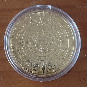 May include: A gold-colored coin with a circular design featuring a central image of a face surrounded by intricate patterns and symbols. The coin is encased in a clear plastic holder.