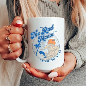 May include: White ceramic mug with a blue and orange graphic of a wave, palm tree, and dolphin. The text "The Rad Mama Catch The Waves" is printed on the mug.
