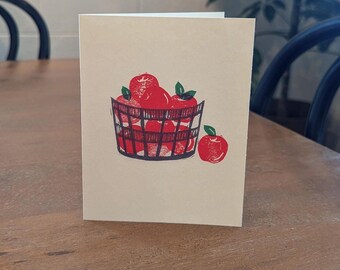 May include: A white card with a red and green illustration of a basket full of apples. The basket is made of woven wood and has a dark brown color. There is a single red apple sitting outside of the basket.