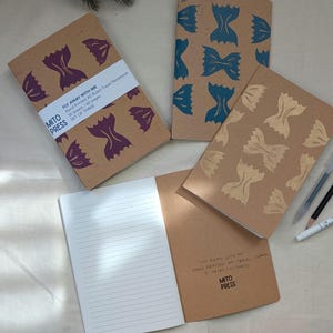 Pode incluir: Conjunto de três cadernos de viagem marrons com um design impresso à mão de um laço. Os cadernos são intitulados "Fly Away With Me" e "Hand Printed AS Ruled Travel Journal".