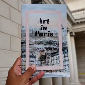 May include: A person holding a travel zine titled "Art in Paris." The cover features a watercolor illustration of Parisian rooftops and the text "AN ART AND TRAVEL ZINE." The zine has a pink border with additional text.