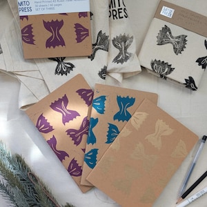 Pode incluir: Conjunto de três cadernos de viagem impressos à mão com capa castanha e um padrão repetitivo de formas de massa de cor roxa, turquesa e dourada. Os cadernos são intitulados "Fly Away With Me" e dizem "Caderno de viagem A5 com régua impresso à mão, 30 folhas/60 páginas, Conjunto de três".