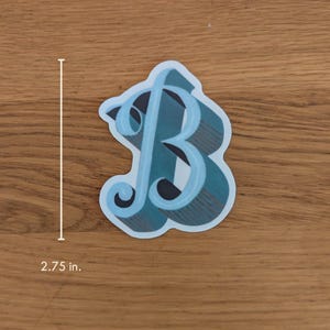 May include: A sticker featuring a stylized letter "B" in shades of teal and blue, with a plaid pattern. The sticker is approximately 2.75 inches in height. The sticker is on a wooden surface.