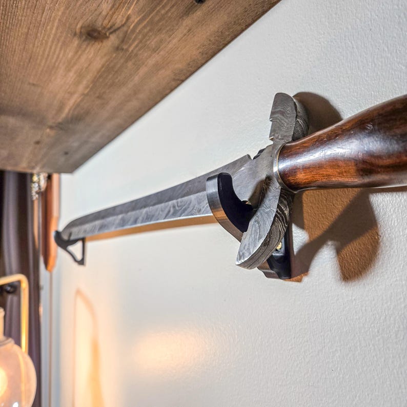 A Damascus steel sword with a wooden handle displayed horizontally on a wall using black sword wall mounts. The brackets securely cradle the blade beneath a rustic wooden shelf, turning the sword into a striking decorative centerpiece for collectors and home décor.