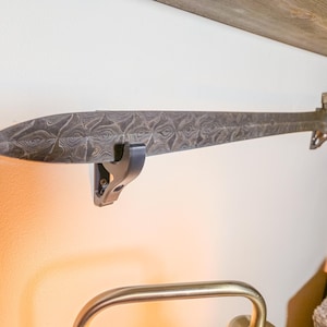 A Damascus sword elegantly displayed horizontally on a wall, securely held by two sleek black hook hangers. The intricate wavy patterns of the Damascus steel blade catch the light, making the sword a striking centerpiece, perfect for collectors and enthusiasts."