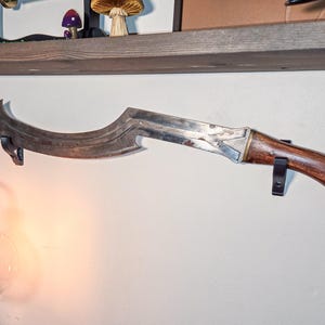 A curved ancient Egyptian khopesh sword with a wooden handle displayed horizontally on a wall using black sword wall mounts. The secure brackets support the blade beneath a rustic wooden shelf, with a glowing lamp nearby, creating a warm and stylish decorative display for sword collectors.