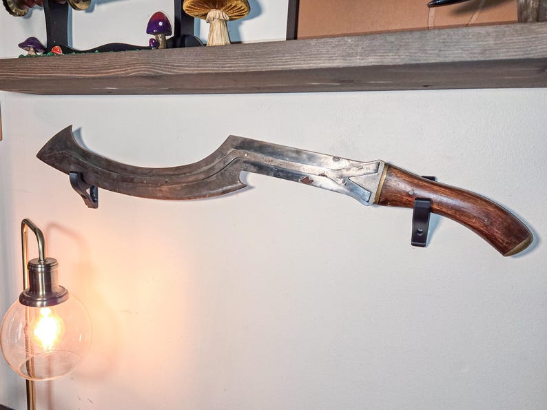 A curved ancient Egyptian khopesh sword with a wooden handle displayed horizontally on a wall using black sword wall mounts. The secure brackets support the blade beneath a rustic wooden shelf, with a glowing lamp nearby, creating a warm and stylish decorative display for sword collectors.