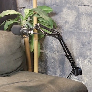 May include: A black microphone arm mounted to a wall with a microphone attached. The arm is adjustable and can be positioned in a variety of ways. The microphone is a condenser microphone with a black finish.