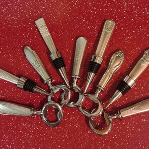 Peut inclure: Un ensemble de huit ouvre-bouteilles en argent avec des poignées ornées. Les ouvre-bouteilles sont disposés en éventail sur une surface rouge mouchetée.