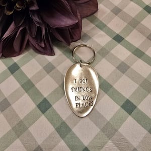 Puede incluir: Un llavero de cuchara de plata con el texto "I got friends in low places" grabado en él. El llavero está sobre una superficie a cuadros blancos y verdes.