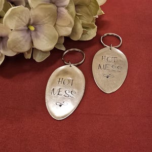 Puede incluir: Dos llaveros plateados con las palabras "HOT MESS" grabadas. Cada llavero tiene un pequeño diseño de abeja. Los llaveros tienen forma ovalada y están unidos a un llavero plateado. El fondo es de tela roja oscura.