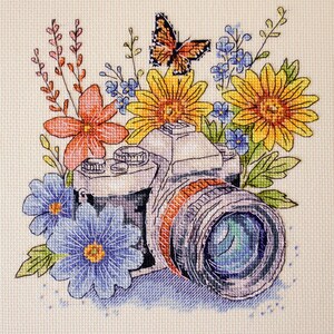 May include: Cross-stitch design featuring a camera surrounded by colorful flowers and a butterfly. The flowers include yellow sunflowers, blue and orange blooms, and green leaves. The camera lens has a blue and green hue.