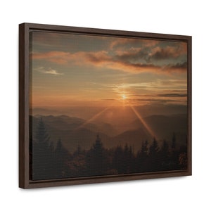 May include: A panoramic view of a mountain range at sunset. The sun is setting behind the mountains, casting a warm glow over the landscape. The sky is a mix of orange, pink, and purple, and the clouds are wispy and white. The mountains are covered in trees, and the air is hazy.