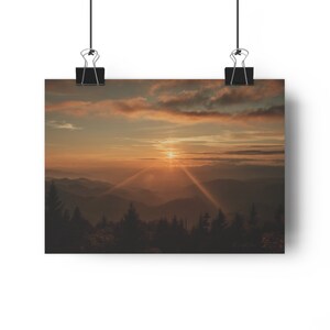 May include: A panoramic view of a mountain range at sunset, with the sun shining through the clouds and casting long rays of light over the landscape. The mountains are covered in trees and the sky is a mix of orange, pink, and purple.