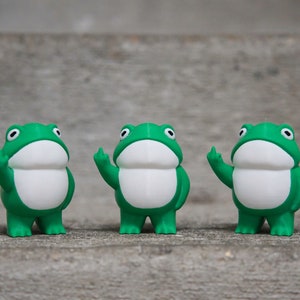 Peut inclure: Trois figurines de grenouilles vertes avec des ventres blancs et des bras lev&eacute;s, chaque doigt &eacute;tant &eacute;tendu, debout sur une surface grise.