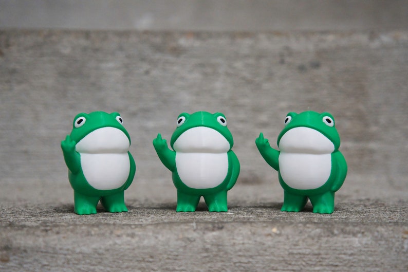 Peut inclure: Trois figurines de grenouilles vertes avec des ventres blancs et des bras lev&eacute;s, chaque doigt &eacute;tant &eacute;tendu, debout sur une surface grise.