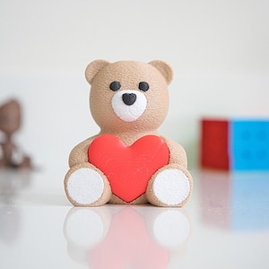 May include: A brown teddy bear holding a red heart in its paws. The bear has a white nose and white paws.