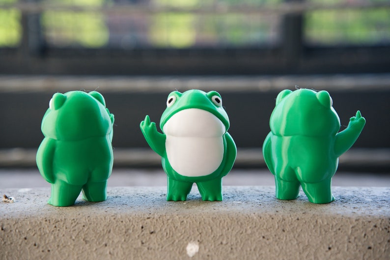 Peut inclure: Trois figurines de grenouilles vertes, chacune avec une pose diff&eacute;rente. La grenouille du milieu fait un pouce en l'air, tandis que les deux autres grenouilles sont debout, les bras le long du corps.