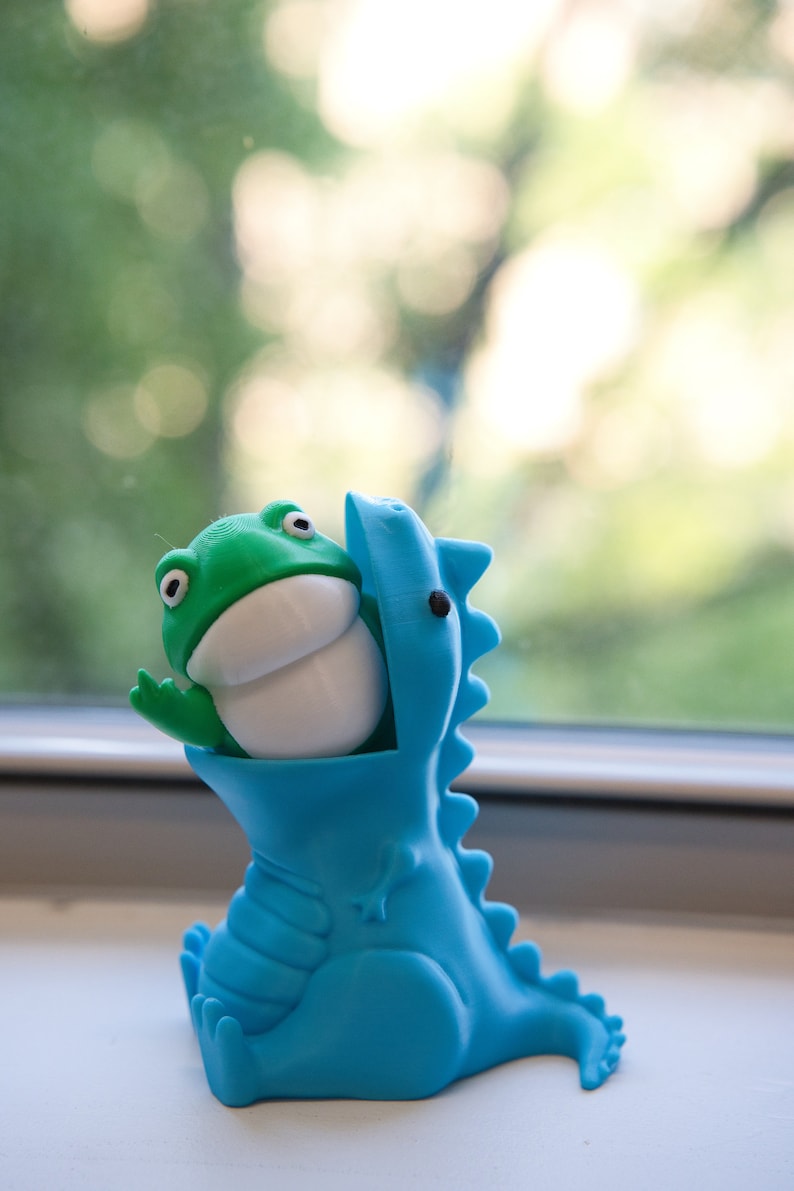 Peut inclure: Un jouet dinosaure bleu avec un jouet grenouille vert dans sa bouche. Le dinosaure est assis sur une surface blanche.