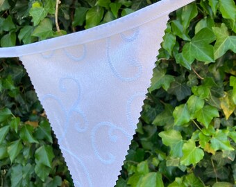 May include: White fabric bunting with a scalloped edge and a delicate swirl pattern. The bunting is hanging against a backdrop of green leaves.