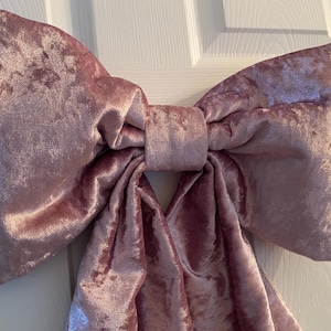 Op de afbeelding: Een grote, pluche strik in een stoffige roze kleur. De strik is gemaakt van een zachte, fluwelen stof met een lichte glans. De strik is in het midden geknoopt en heeft lange, vloeiende staarten. De strik wordt waarschijnlijk gebruikt voor decoratie.