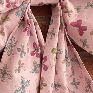 May include: A pink fabric with a butterfly pattern. The butterflies are in shades of pink, brown, and gray, with delicate details. The fabric is gathered and folded, creating a bow-like shape. The background is a brick wall.