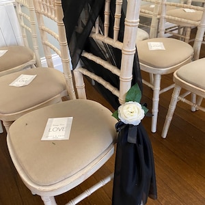 May include: Cream-colored wooden chairs with tan cushions are decorated with black fabric and white roses. The chairs have a classic design, and some have small cards that say "LOVE" on the seat.