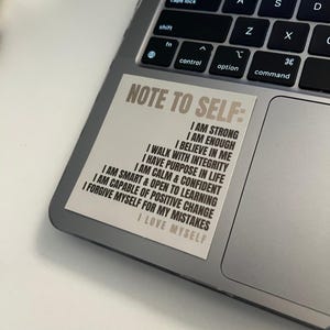 Pu&ograve; includere: Un adesivo beige con il testo "Note to Self:" e un elenco di affermazioni positive in testo nero. L'adesivo &egrave; su un laptop argentato.